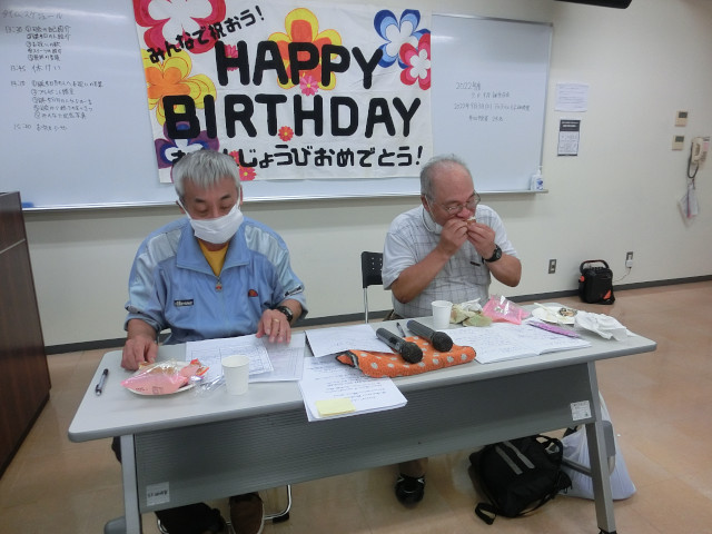 司会している2名の男性メンバー。机に座っている。机の上には司会者用のプログラム。そしてお菓子がある。どちらも白髪で高齢。うち1人は司会をしているにも関わらずお菓子にかぶりついている。