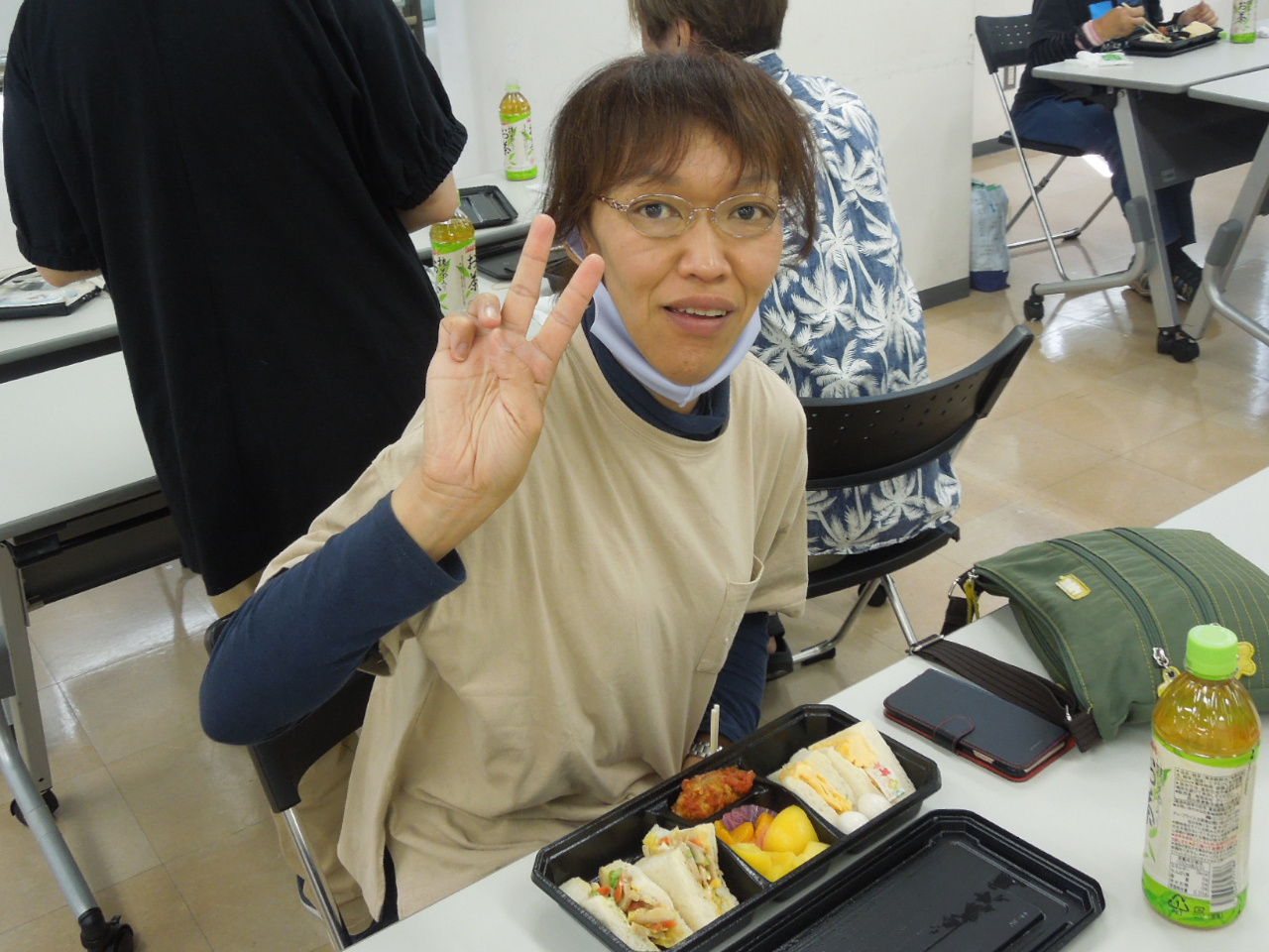 誕生日会に参加している女性メンバーの写真。机に座っていて、目の前には食べかけのサンドイッチがある。ピースをしている。