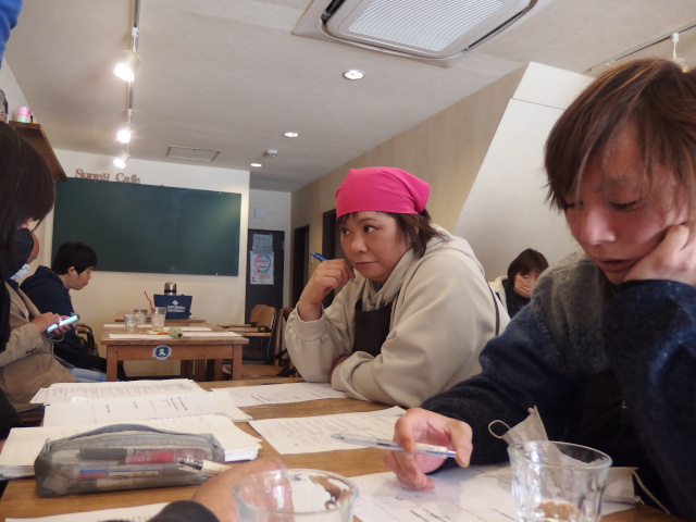 学習会に参加する男女。スタッフ1名が司会をしている。仕事の途中で抜け、学習会に参加したメンバーもいる。その人はエプロン・三角巾を付けている。皆、机に座っている。紙を机に置いて見ている。