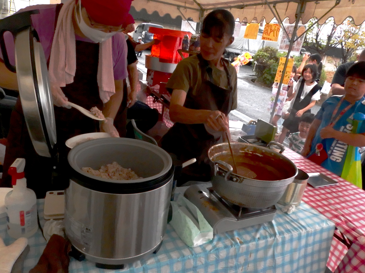 出店でカレーを用意しているメンバー・スタッフの男女2名。男性メンバーがごはんをよそっている。女性スタッフがカレールーをレードルに入れている。お客の子供がカレーを待っている。