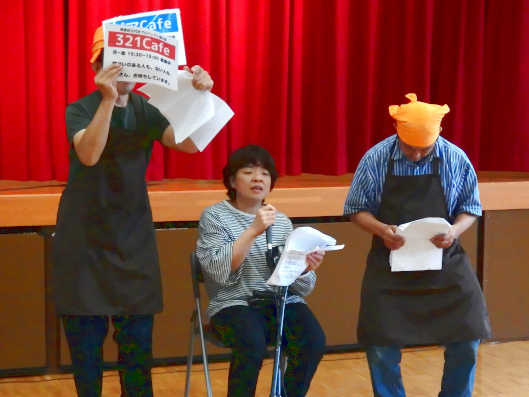 講演をしている男女。スタッフ1名とメンバー2名。スタッフとメンバー1名づつマイクを握って講演中。1人はプラカードを掲げている。