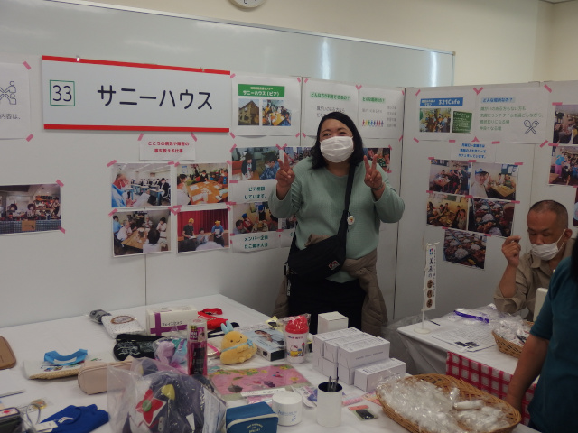 机の上にクイズの景品が並んでいる。女性が机の奥でピースをしている。女性の後ろの壁には活動内容を印刷して張りつけている。