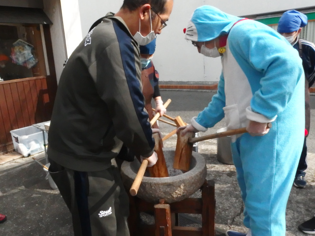 男性3名が餅を杵で潰している。うち1名はドラえもんの着ぐるみを着ている。子供に親しみやすくなるためだ。