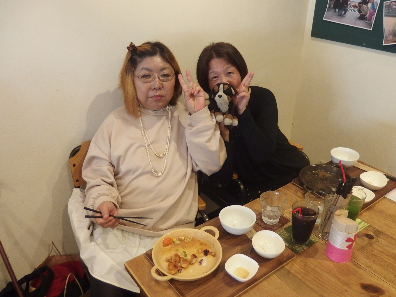 90歳ぐらいの高齢の男性のお客と男性メンバーが写っている写真。場所はカフェのホール。男性メンバーはピースをしている。高齢の男性は机に向かって座っている。机の上にはパウンドケーキとココアがある。