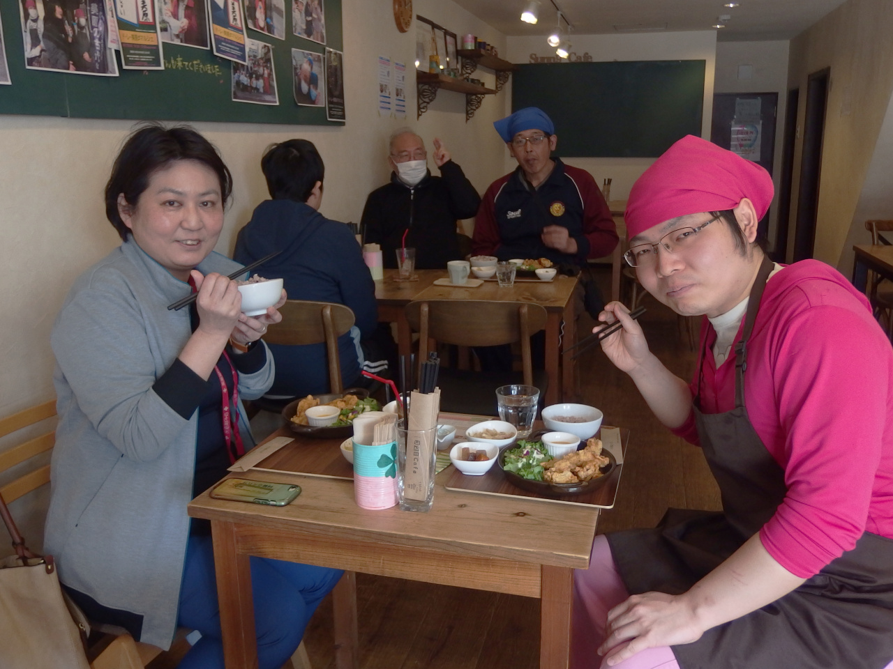 5人の男女が集まっている写真。場所はカフェのホール。手前に写っている女性のお客とメンバーの男性がご飯を食べている。2人とも微笑んでいる。奥の方でも1人ご飯を食べている男性メンバーがいる。他2名はご飯を食べず、机に向かっている。1人高齢の男性メンバーがピースしている。