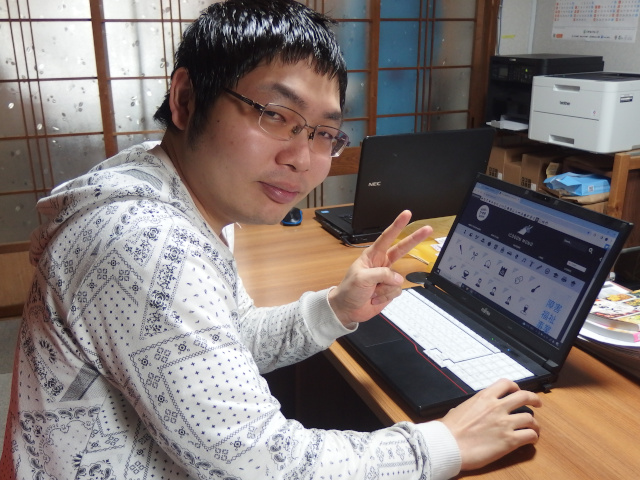 PC作業をする男性メンバーの写真。こちらに向かってピースをしている。