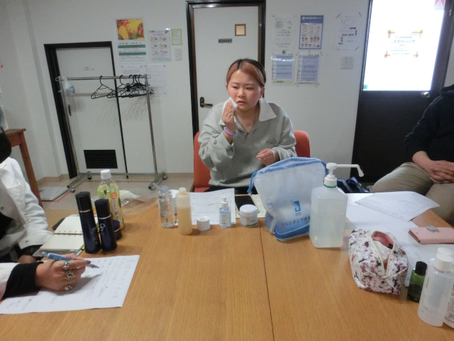 化粧をしているメンバー女性の写真。化粧水を肌に塗りこんでいる。椅子に座って机に向かっている。机には化粧品が沢山置いてある。写真からは切れてしまっているが周りには人が集まっている。