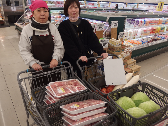 スーパーで買い物をする女性2名。スタッフとメンバー。大量の生肉と野菜をカートの上のカゴに入れている。