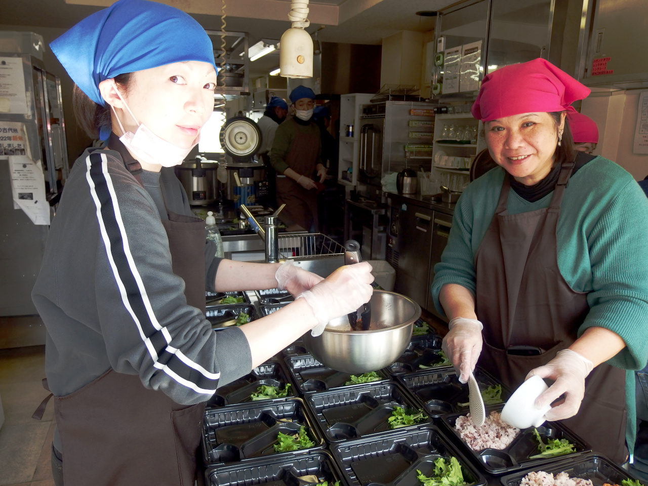 お弁当を作っている女性2人の写真。1人はボールから食材を取り出し、お弁当箱に入れようとしている。1人はご飯をよそっている。ボールから食材を取り出している女性はスタッフ。ご飯をよそっている女性はメンバー。