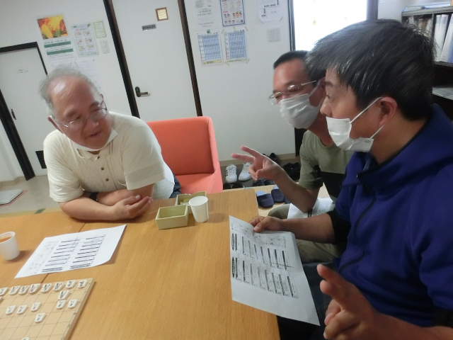 3名の男性メンバー。うち1名が指導者。1名がコマの進め方について質問している。