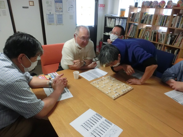 将棋教室の写真。4名の男性メンバーが写っている。椅子に座り、目の前の机に将棋板が置かれている。男性メンバーのうち1人が将棋板を以外そうに覗き込んでいる。彼はリアクションが激しい性格をしているので、恐らく指導を受ける上で以外な事があったのだろう。