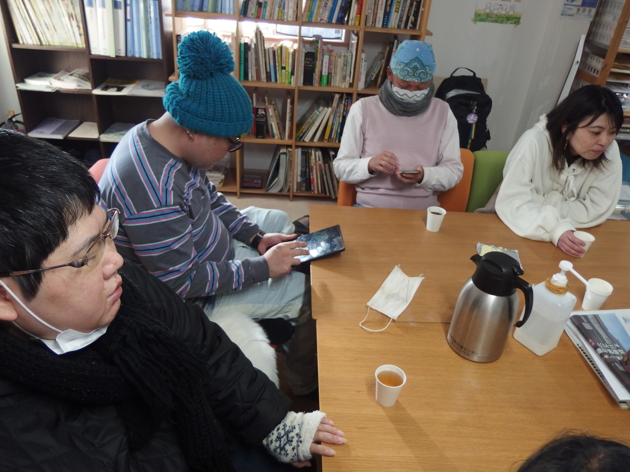 椅子に座り、リラックスしている男女の写真。うち2名メンバー。うち1名スタッフ。うち1名ボランティア。2名がスマホを操作している。1名は話に夢中になっている。1名はとくになにもせず座っている。