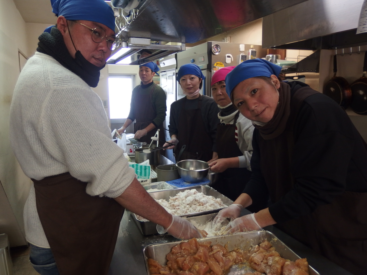 5人ほどの男女が厨房に居る。2人の男性が唐揚げの衣を付けている。調理台の上には調理器具が沢山置いてある。